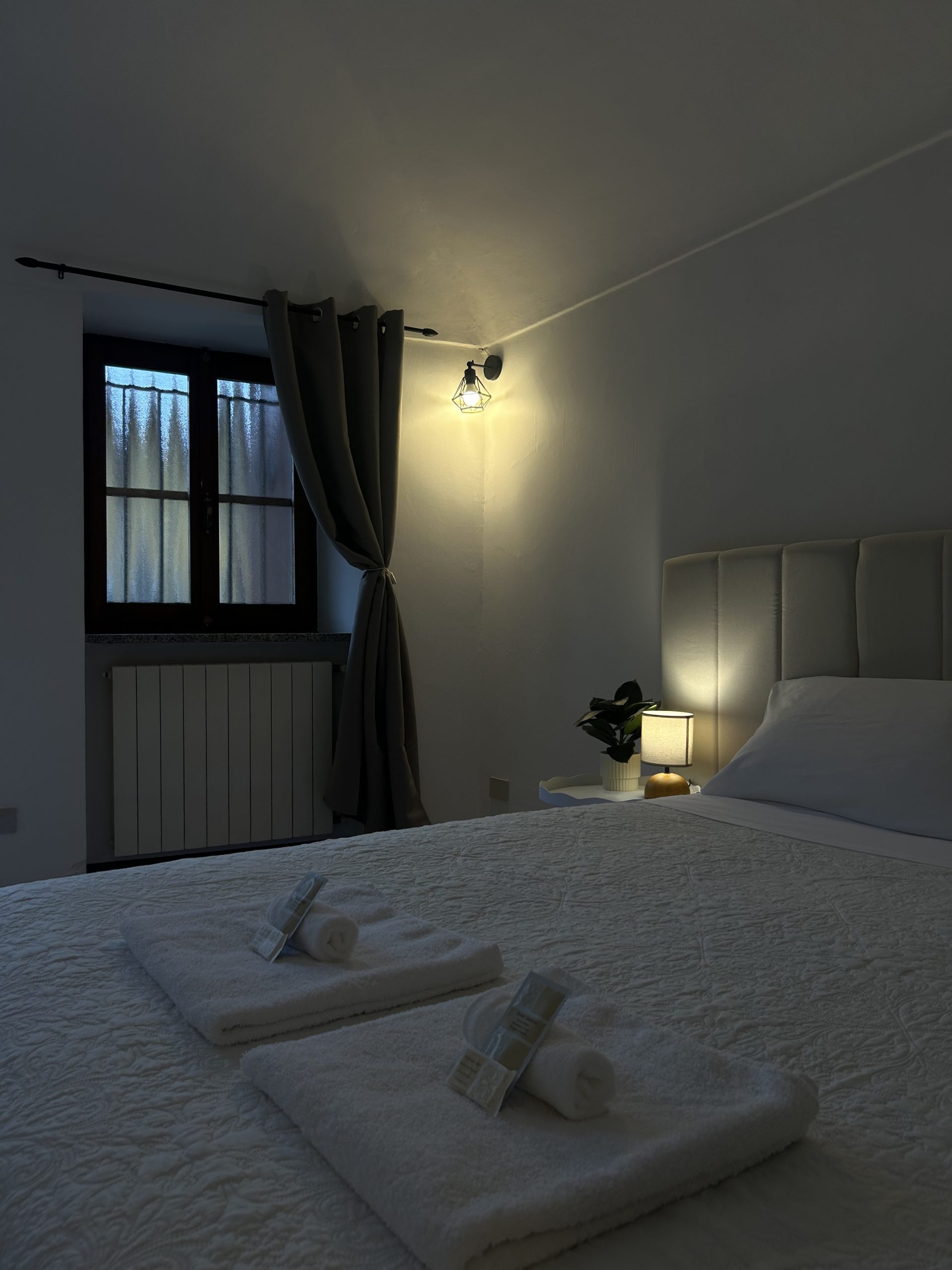 Una camera da letto accogliente con un letto ben rifatto, su cui sono disposti due asciugamani bianchi e alcuni articoli da bagno, una piccola lampada sul comodino e una finestra con tende scure parzialmente chiuse. La stanza è illuminata da una luce soffusa.